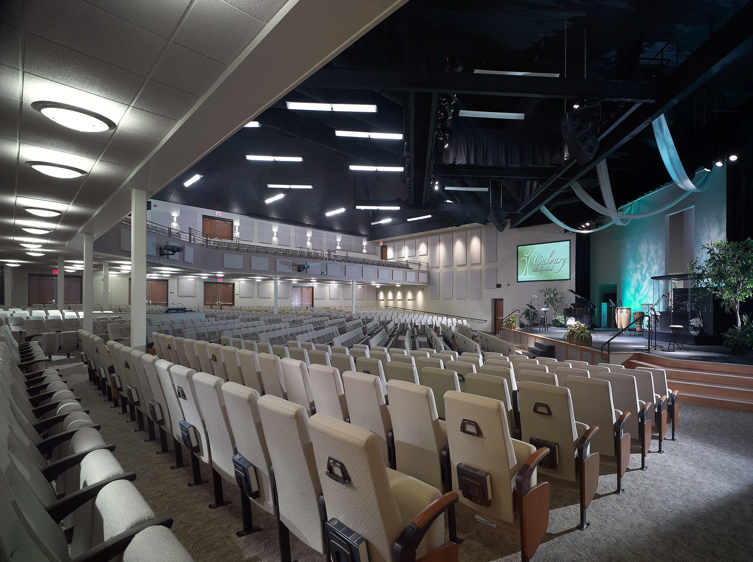 Empty church auditorium with beige cushioned seats, stage with musical instruments, and a large screen displaying 'Calvary'.