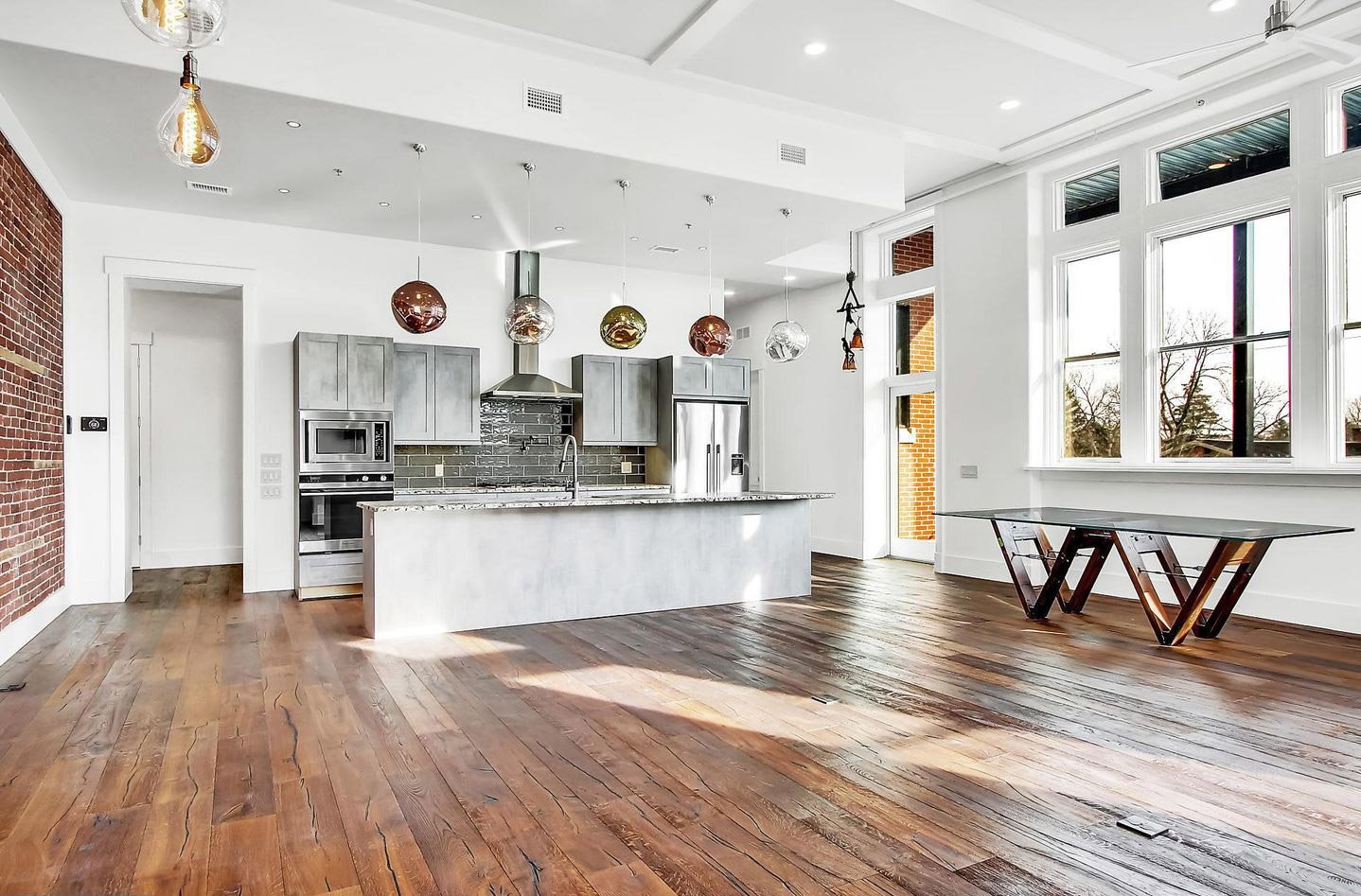Open-concept modern kitchen and dining area with large windows, wooden floors, gray cabinets, a marble kitchen island, hanging glass pendant lights, and a unique glass table with wooden base.