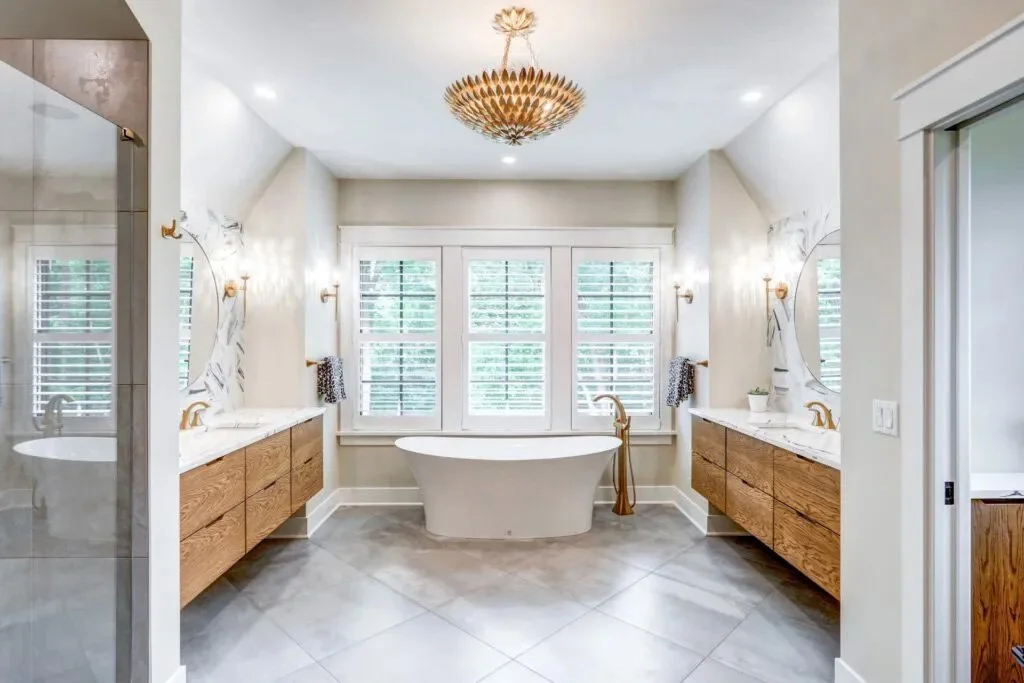 A spacious bathroom with a freestanding bathtub in front of three large windows with white shutters. There are two wooden vanities with white countertops, each with a round mirror, wall-mounted lights, and decorative towels. The floor is tiled, and t