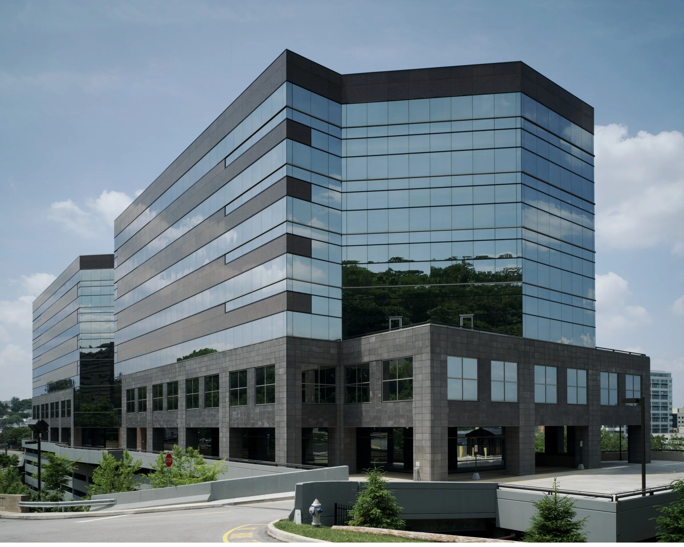 A modern multi-story office building with a glass and concrete facade, reflecting the sky and surrounding trees. The building has horizontal lines and large windows, with a street and small landscaped area in the foreground.