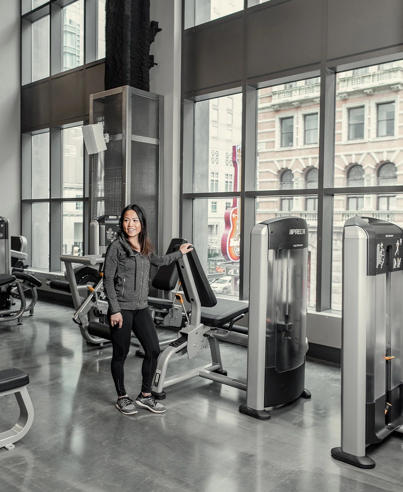 A woman in workout clothes standing next to exercise equipment in a gym with large windows and a city view.