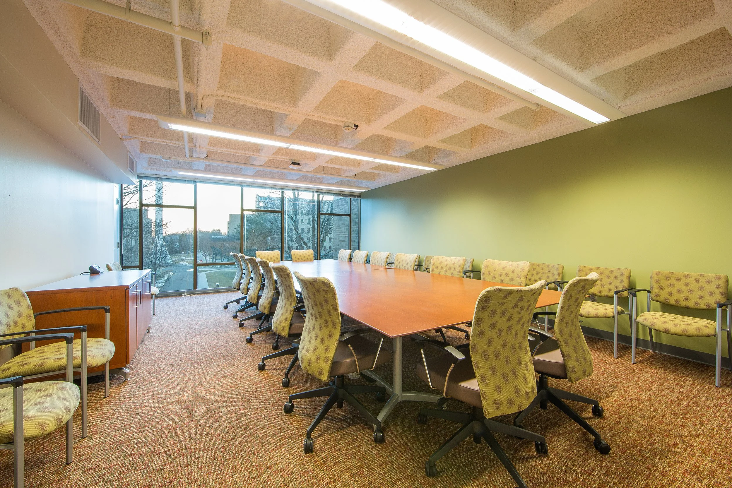 Empty college meeting room with a large wooden table, green patterned chairs, and floor-to-ceiling windows showing trees outside.