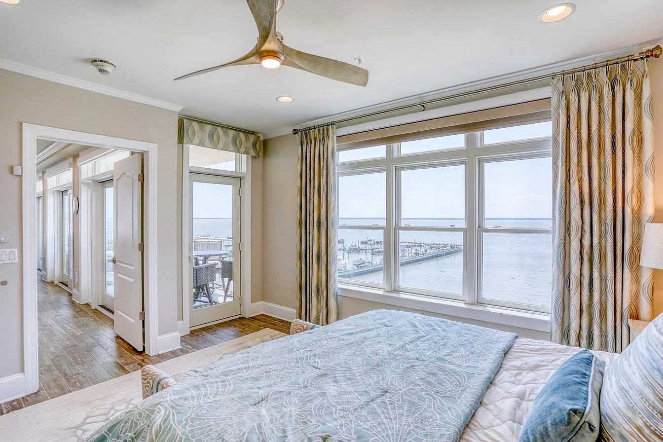 Bedroom with large windows overlooking a marina, bed with patterned bedspread, curtains, ceiling fan, and a door leading to a balcony.