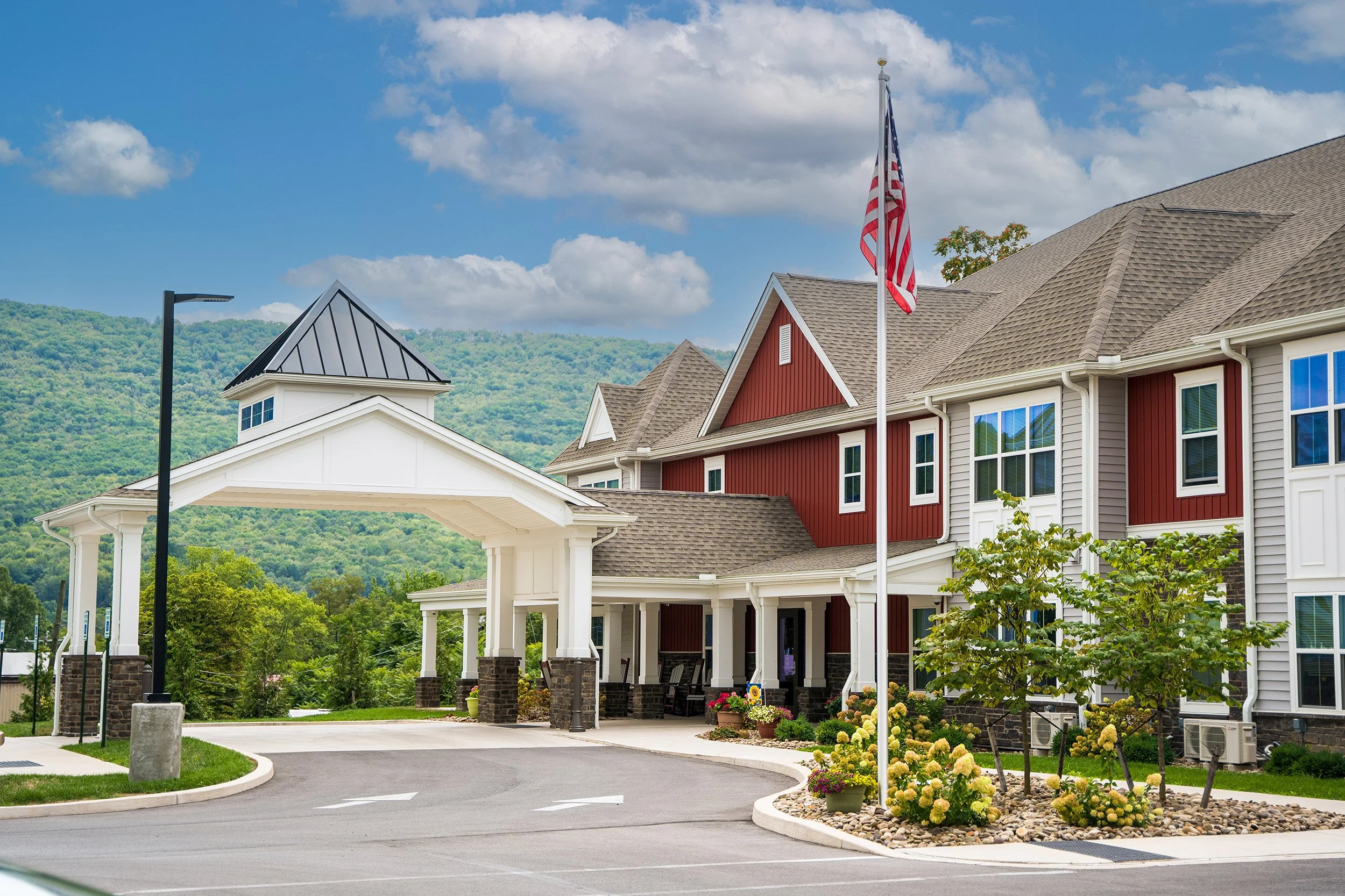 A modern multifamily residential building with a covered entrance, red and gray siding, and a flagpole with an American flag in front of a lush green hill.