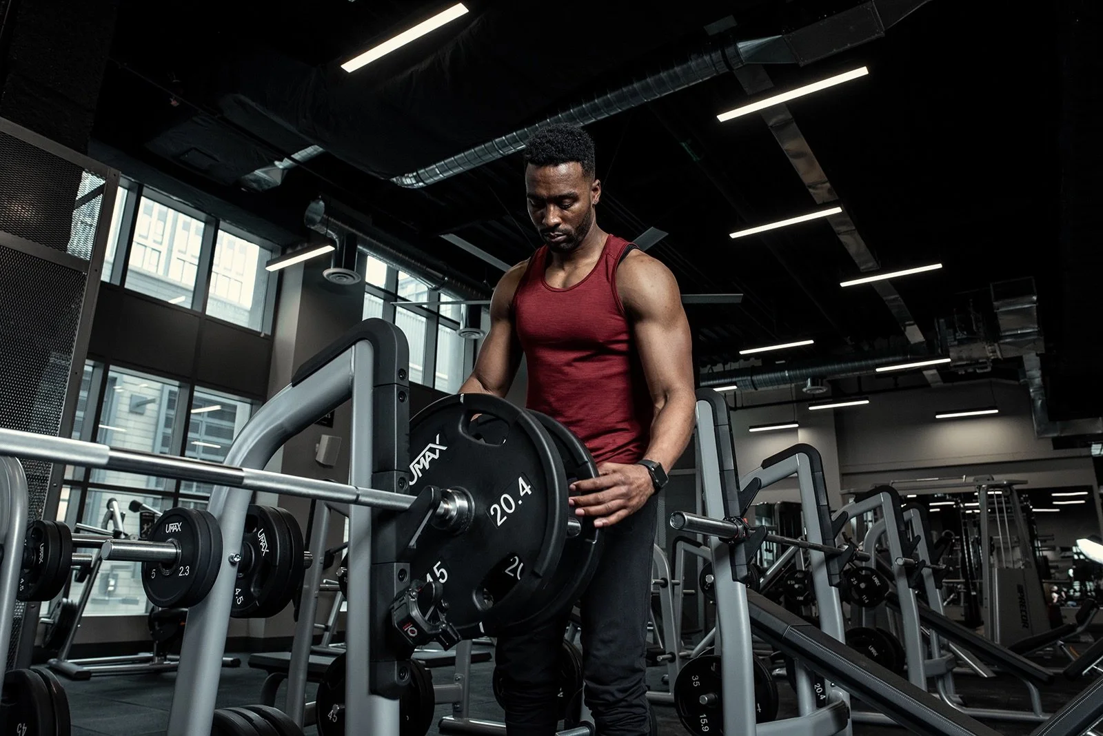 Man lifting a weight plate at a gym with modern equipment and large windows.