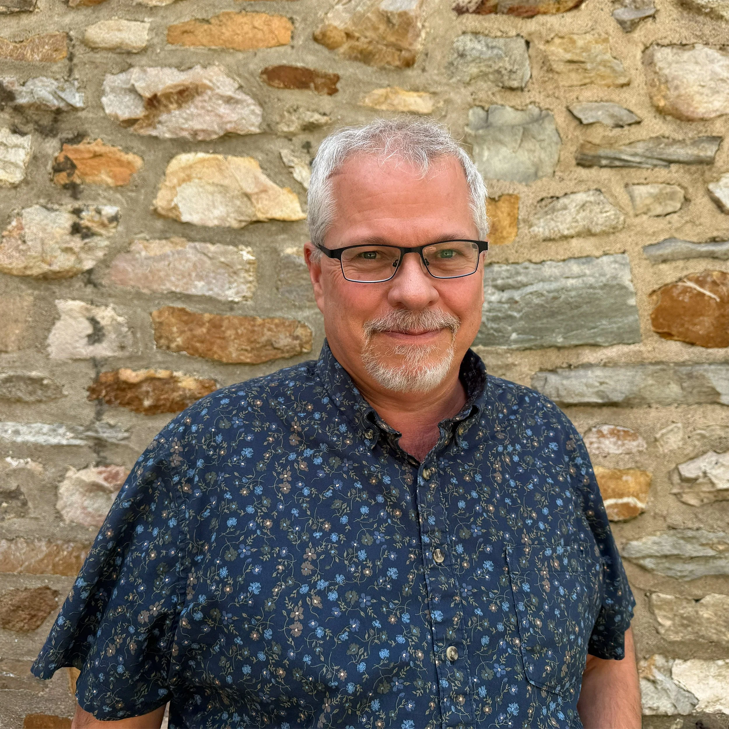 A middle-aged man with gray hair, glasses, and a gray beard wearing a dark blue shirt with a floral pattern, standing in front of a rustic stone wall.