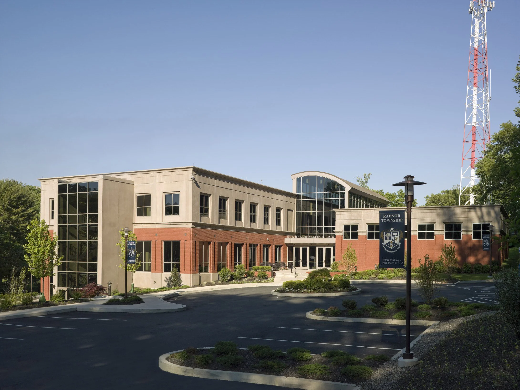 Exterior view of a modern municipal building with brick and concrete facade, large glass windows, surrounded by a parking lot and greenery, with a red and white communication tower nearby.
