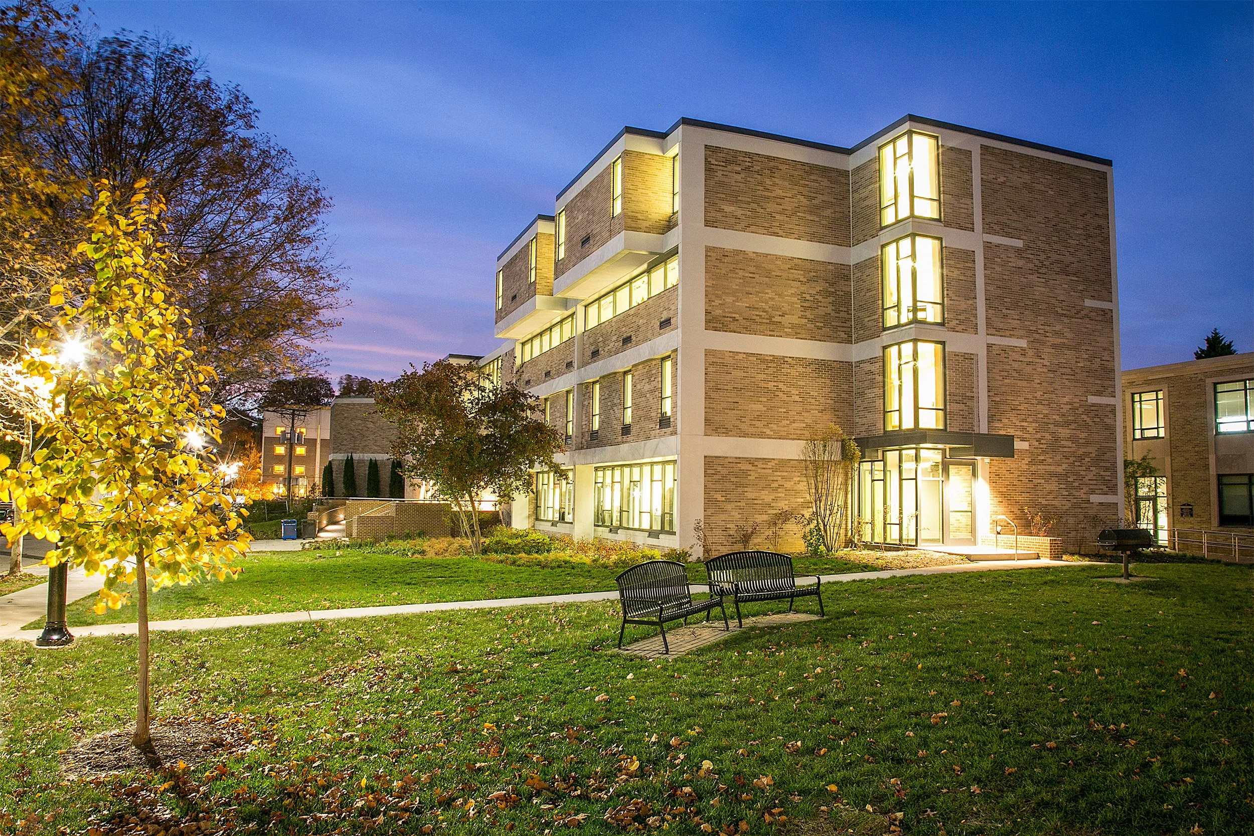A modern multi-story brick and concrete academic building illuminated at dusk, with lit windows, surrounded by trees, grass, and benches in a landscaped area.