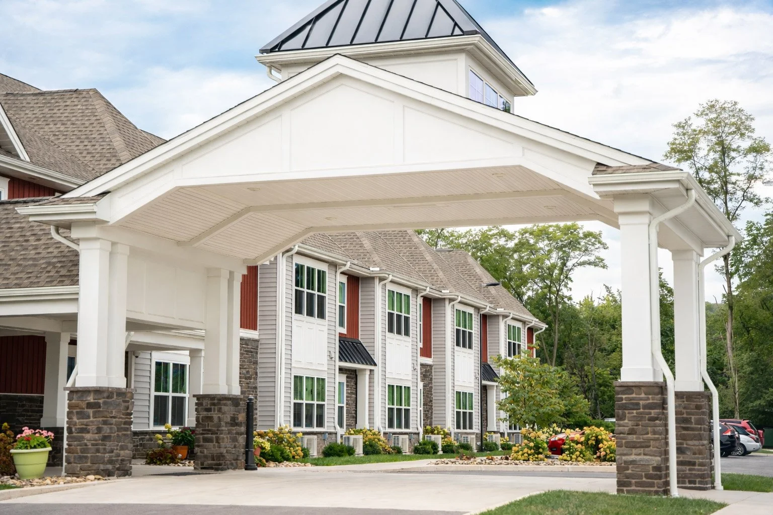 Exterior view of residential building with a covered carport, garden plants, and parked cars, surrounded by trees.