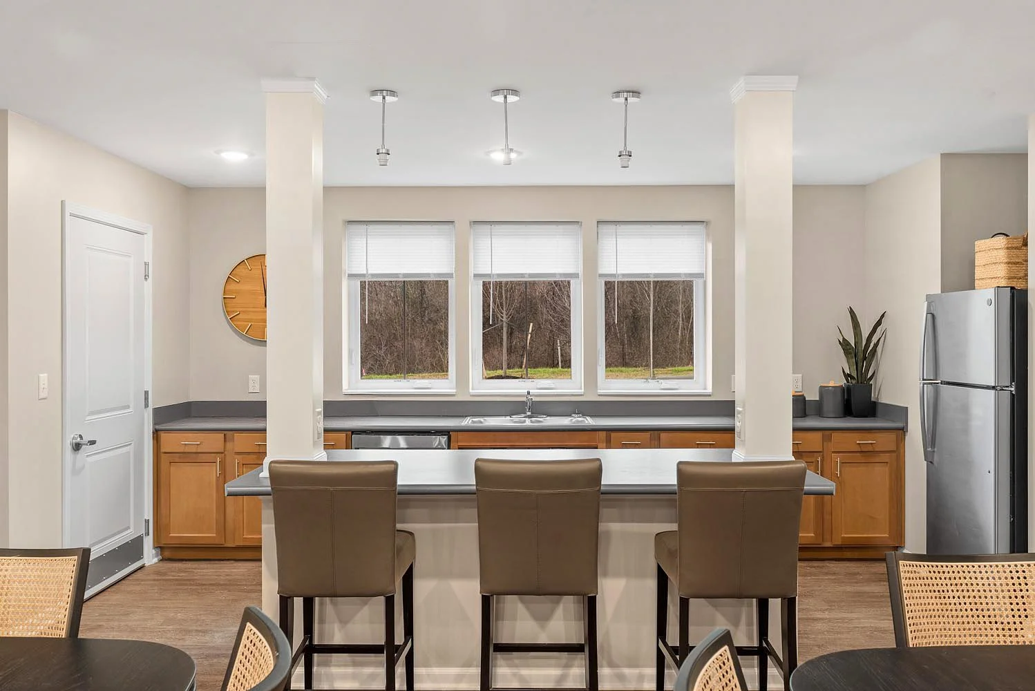 Modern kitchen with three large windows, wooden cabinets, gray countertops, stainless steel refrigerator, ceiling lights, and a breakfast bar with three brown chairs.