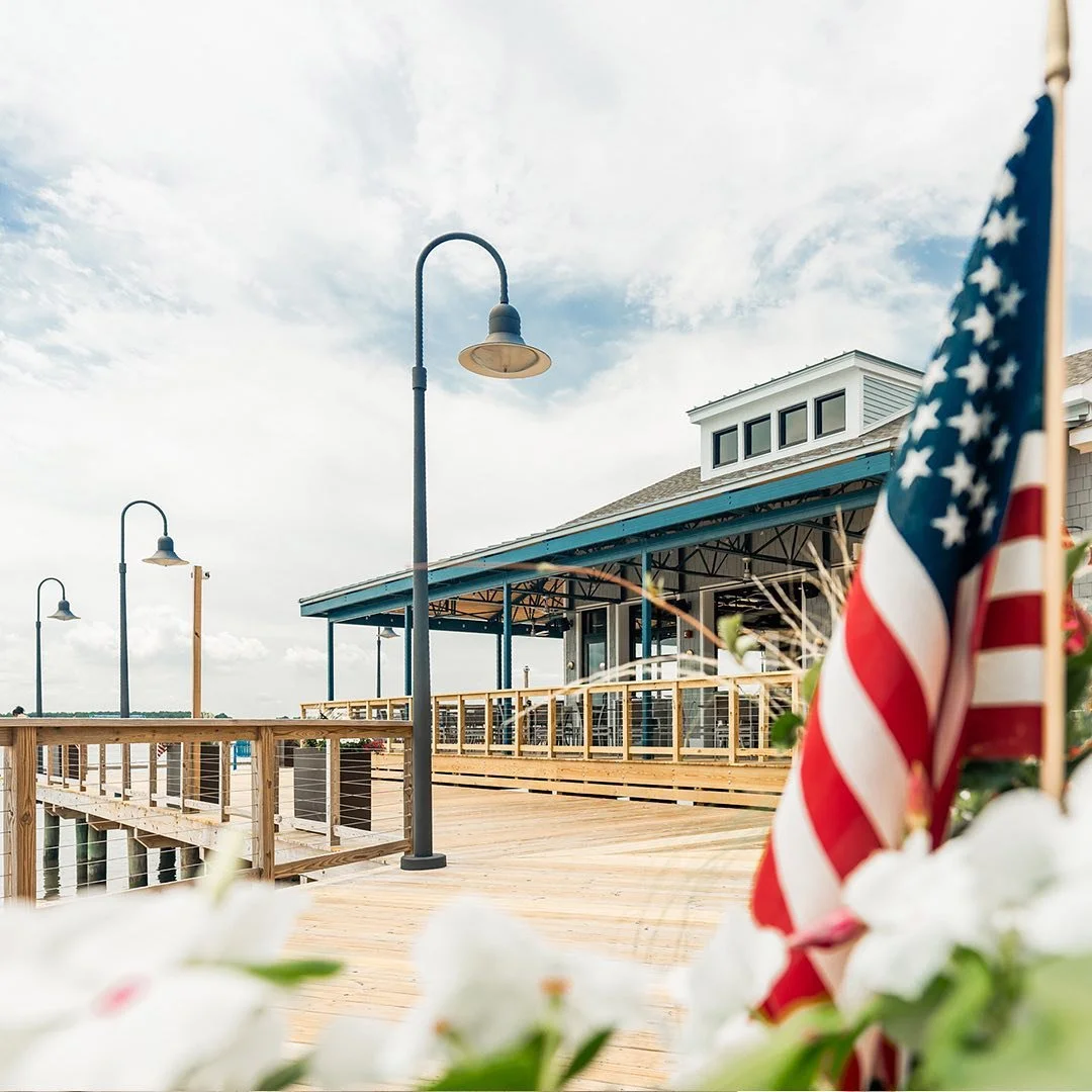 A wooden pier with black street lamps, an American flag, and a building with a covered patio under a cloudy sky.