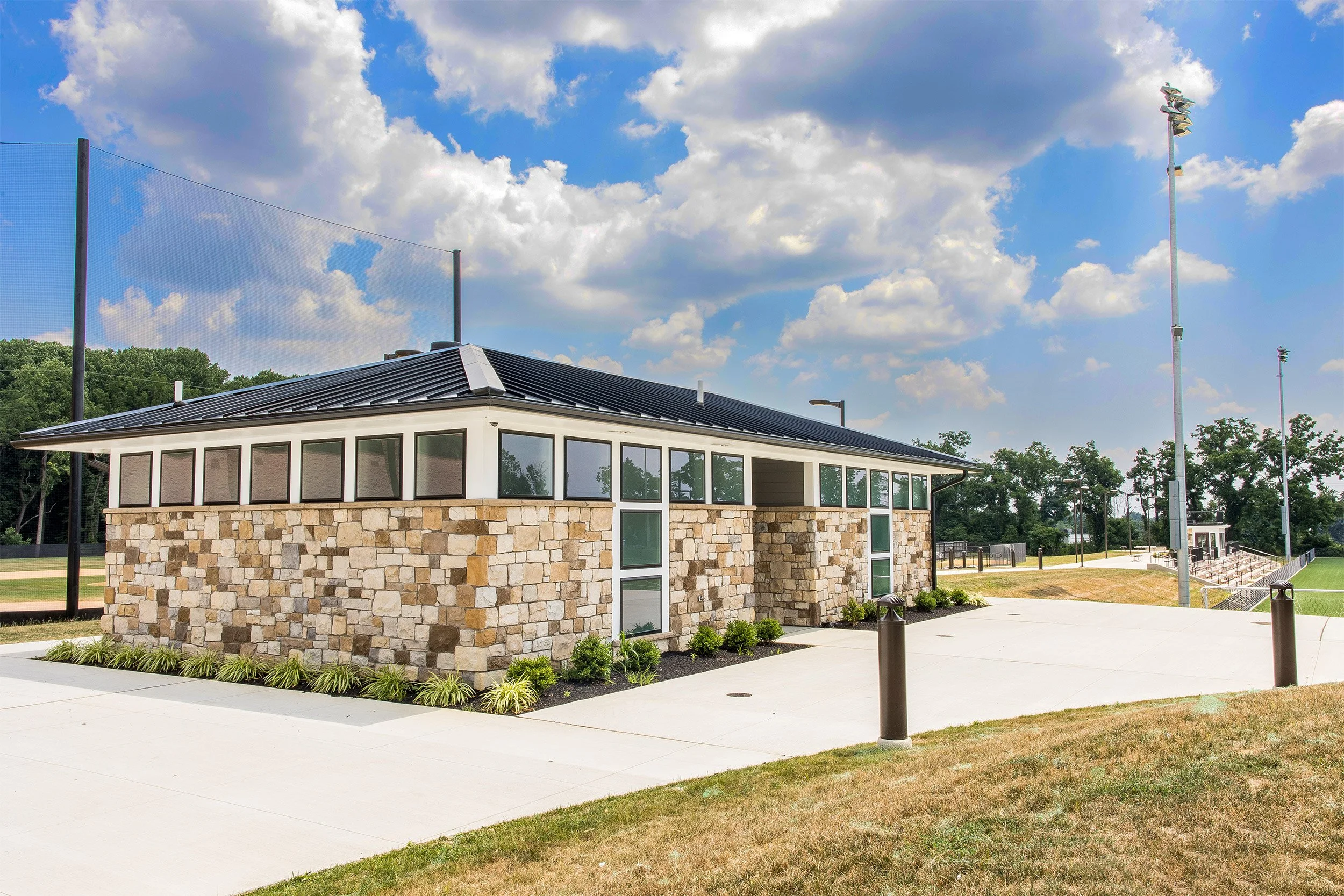 A small restroom facility with stone and white siding, surrounded by a concrete sidewalk, with a sports field and tall light poles in the background under a partly cloudy sky.