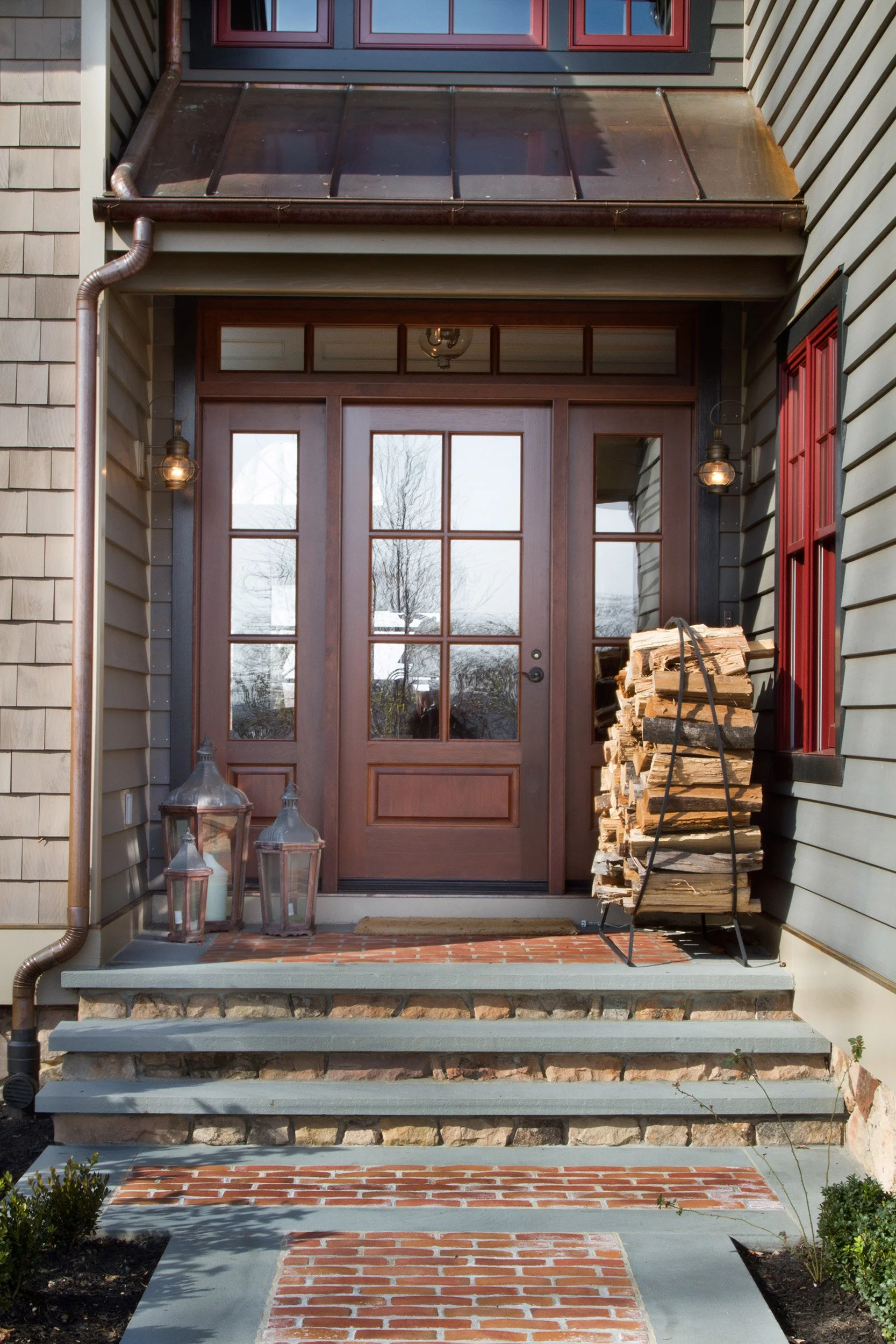 Front porch of a house with a brick pathway, concrete steps, a wooden door with glass panels, lanterns, and a stack of firewood on a metal stand.