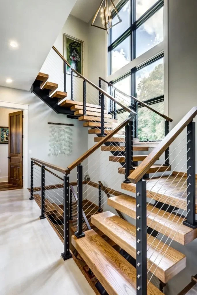Modern staircase with wooden treads, metal supports, and cable railings inside a house with large windows and contemporary decor