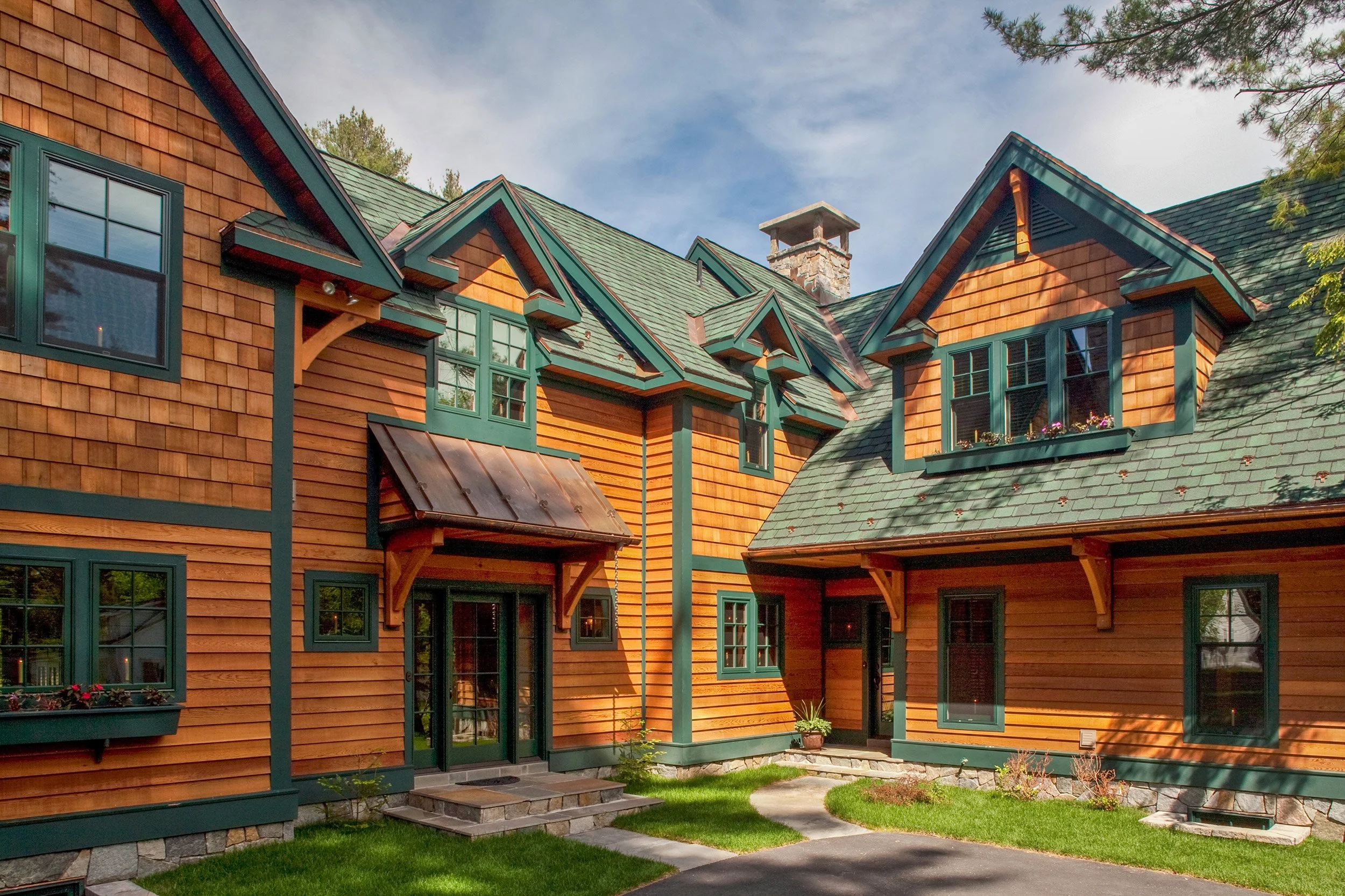 A large, multi-story Adirondack style lakehouse with wooden siding, green window frames, and a green shingled roof, surrounded by a well-maintained lawn and trees.