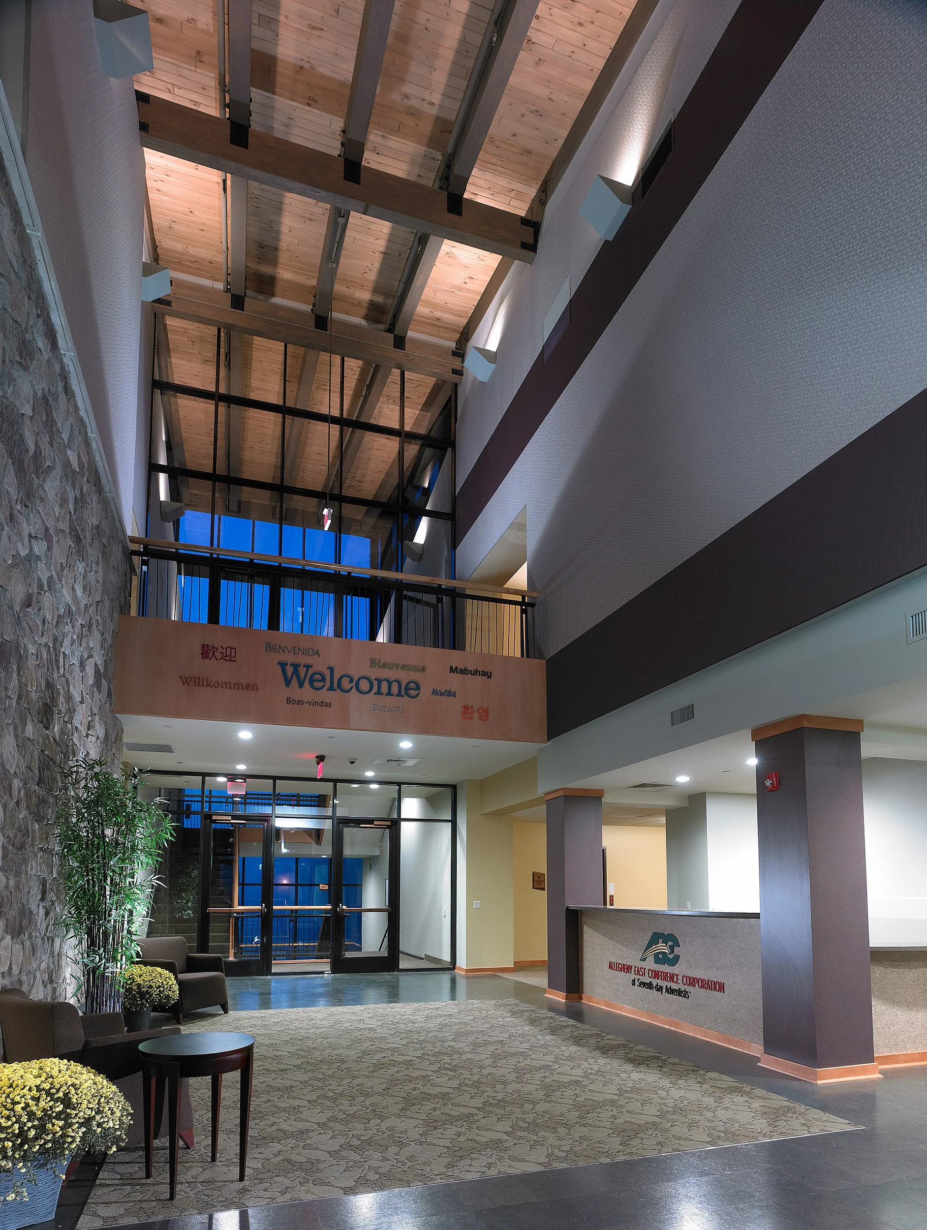 Interior of a modern conference center lobby with stone and drywall walls, a reception desk with a logo, a waiting area with chairs and potted plants, a large window, and a high ceiling with exposed wooden beams and lighting.