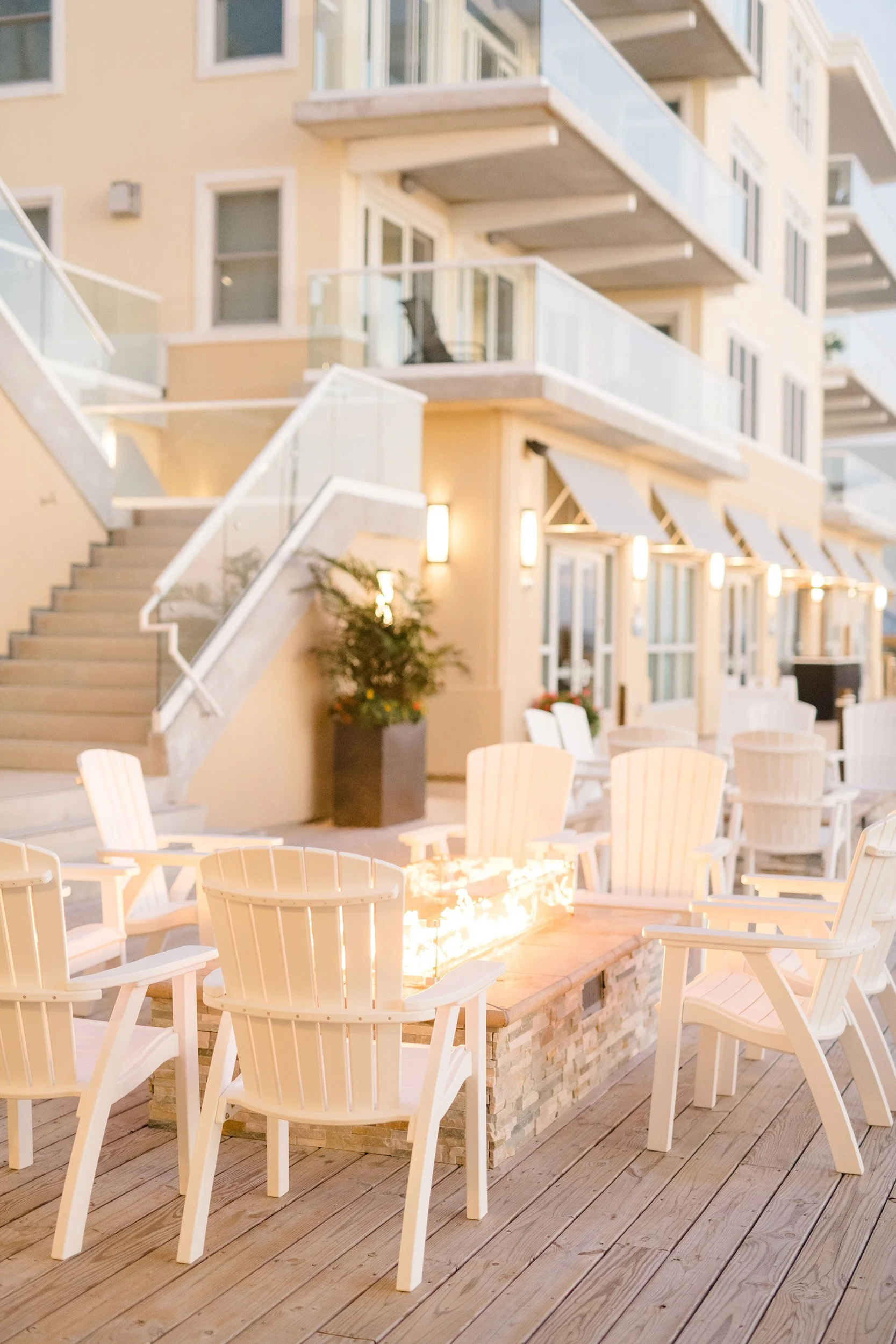 An outdoor patio area with white Adirondack chairs around a rectangular fire pit with a fire. There is a wooden deck floor and a building with multiple small balconies and outdoor lighting fixtures in the background.