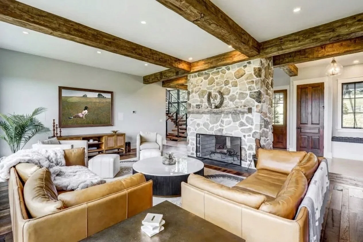 Living room with stone fireplace, wooden ceiling beams, and modern furniture including tan leather couches and white chairs, decorated with artwork and plants.