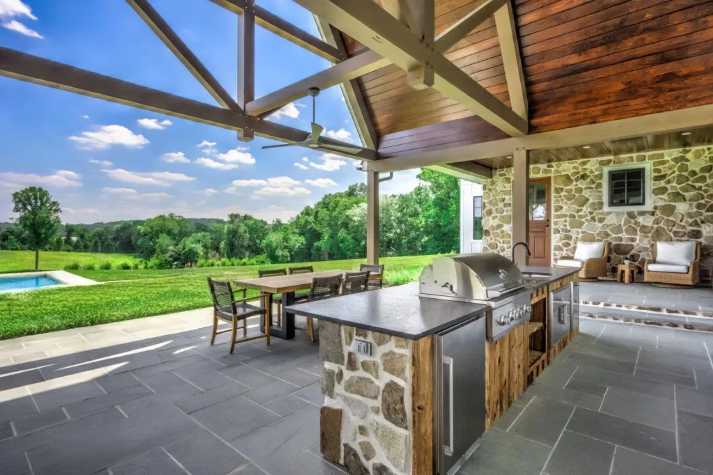 Covered outdoor patio with stone wall backdrop, outdoor kitchen with grill, dining table and chairs, seating area with armchairs, overlooking a lush green landscape and a swimming pool under a partly cloudy sky.