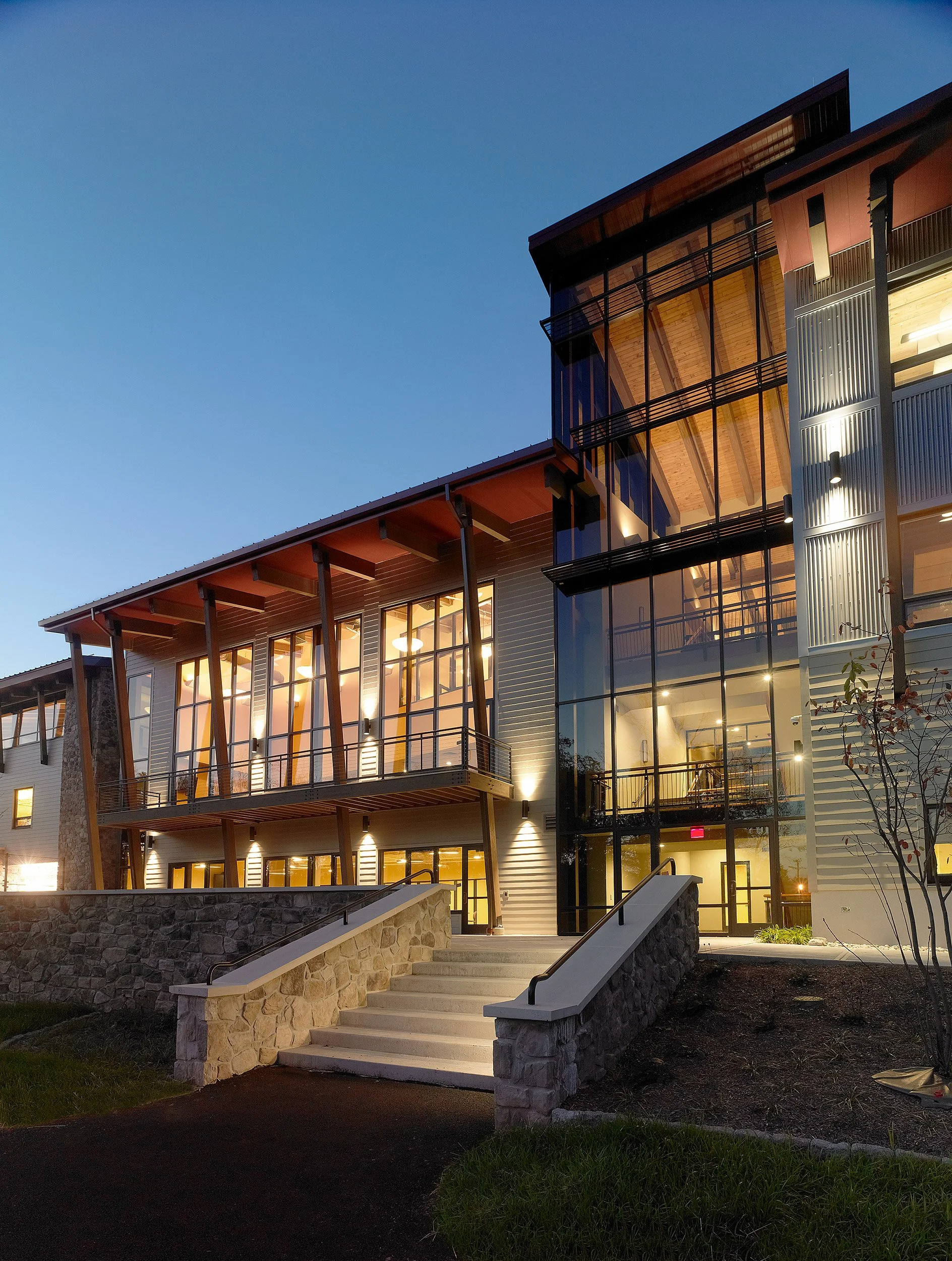 Modern multi-story building with large glass windows and exterior lighting, stone steps leading to entrance, during dusk or early evening.