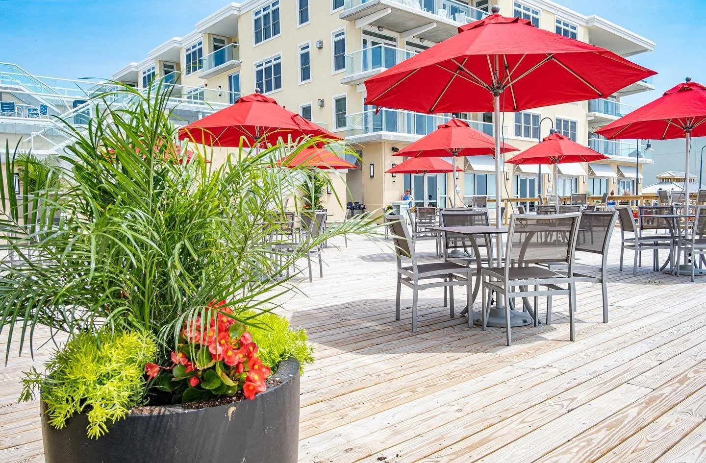 Outdoor patio area with wooden flooring, red umbrellas, grey chairs and tables, large potted green plants, and a multi-story beige building with glass balconies in the background.