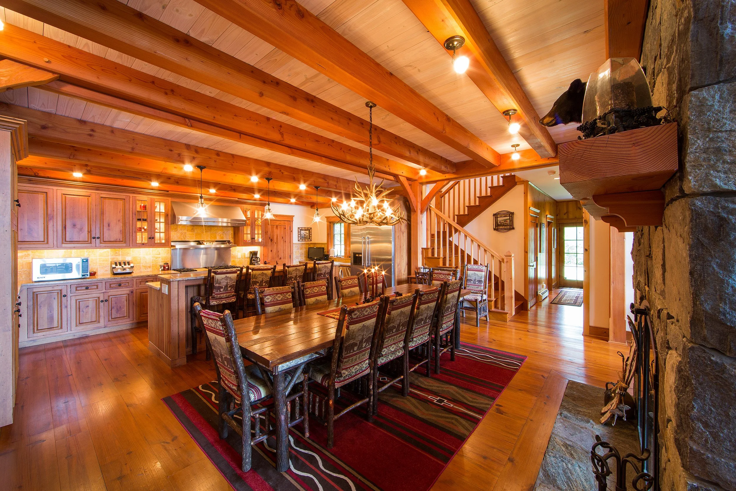 Interior of a rustic Adirondack kitchen and dining area with wooden beams, a long dining table with patterned chairs, a stone fireplace, and a staircase leading upstairs.
