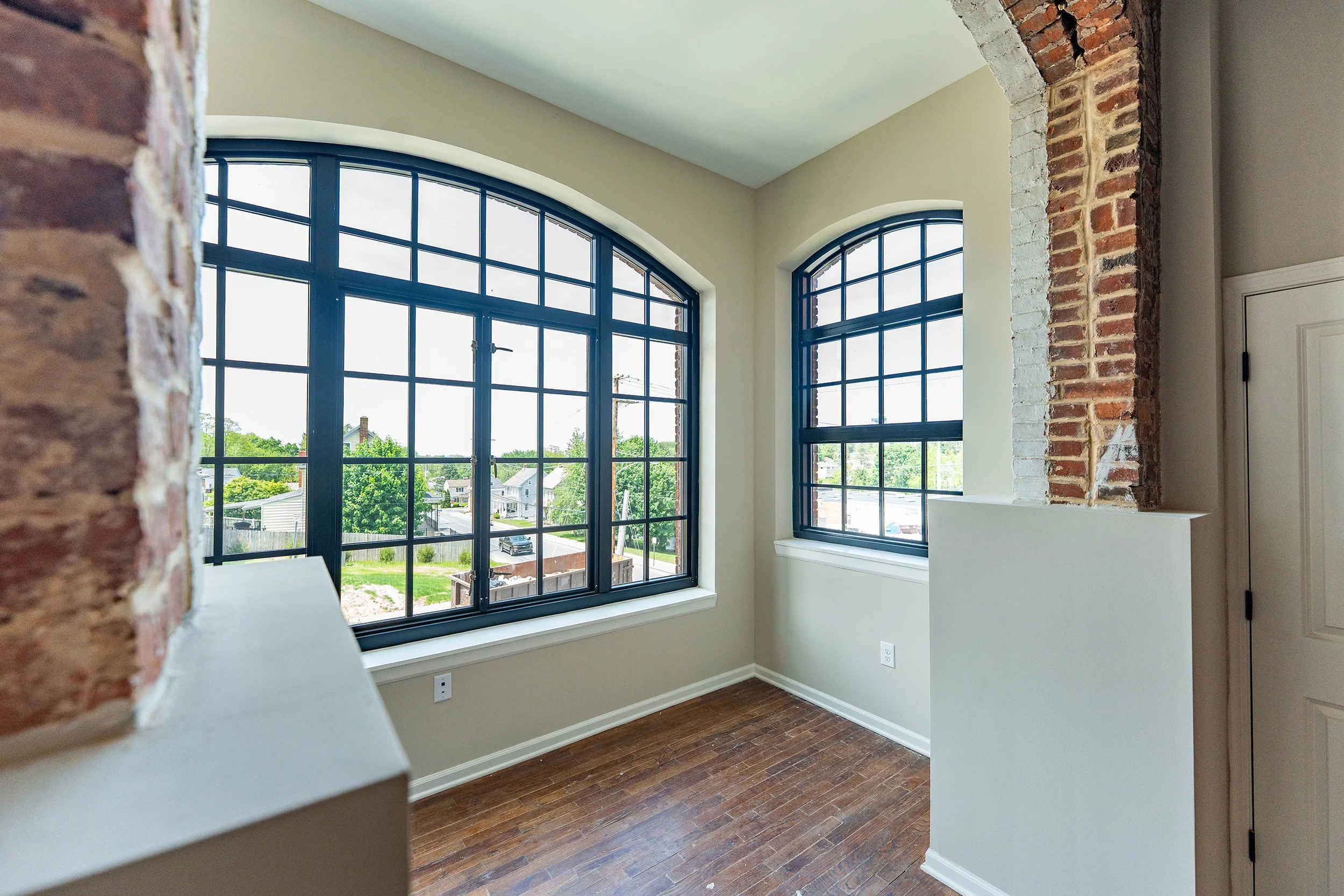 Corner of an apartment with large, arched windows with black frames, exposed brick wall accents, whitewashed trim, light beige walls, and hardwood floors.