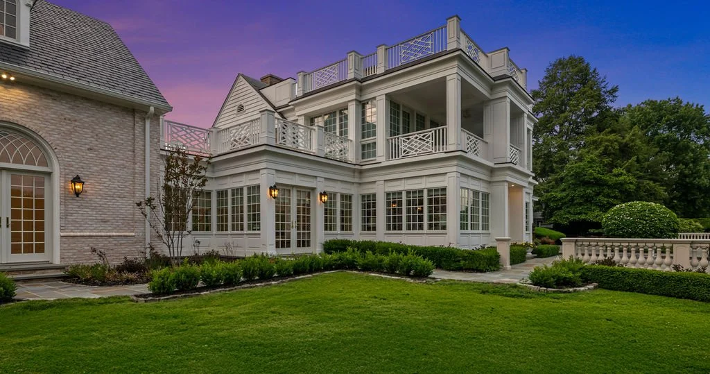 A large, white, multi-story house with large windows, balconies, and a decorative balcony railing, surrounded by a well-maintained lawn and garden, with a purple and pink sky in the background.
