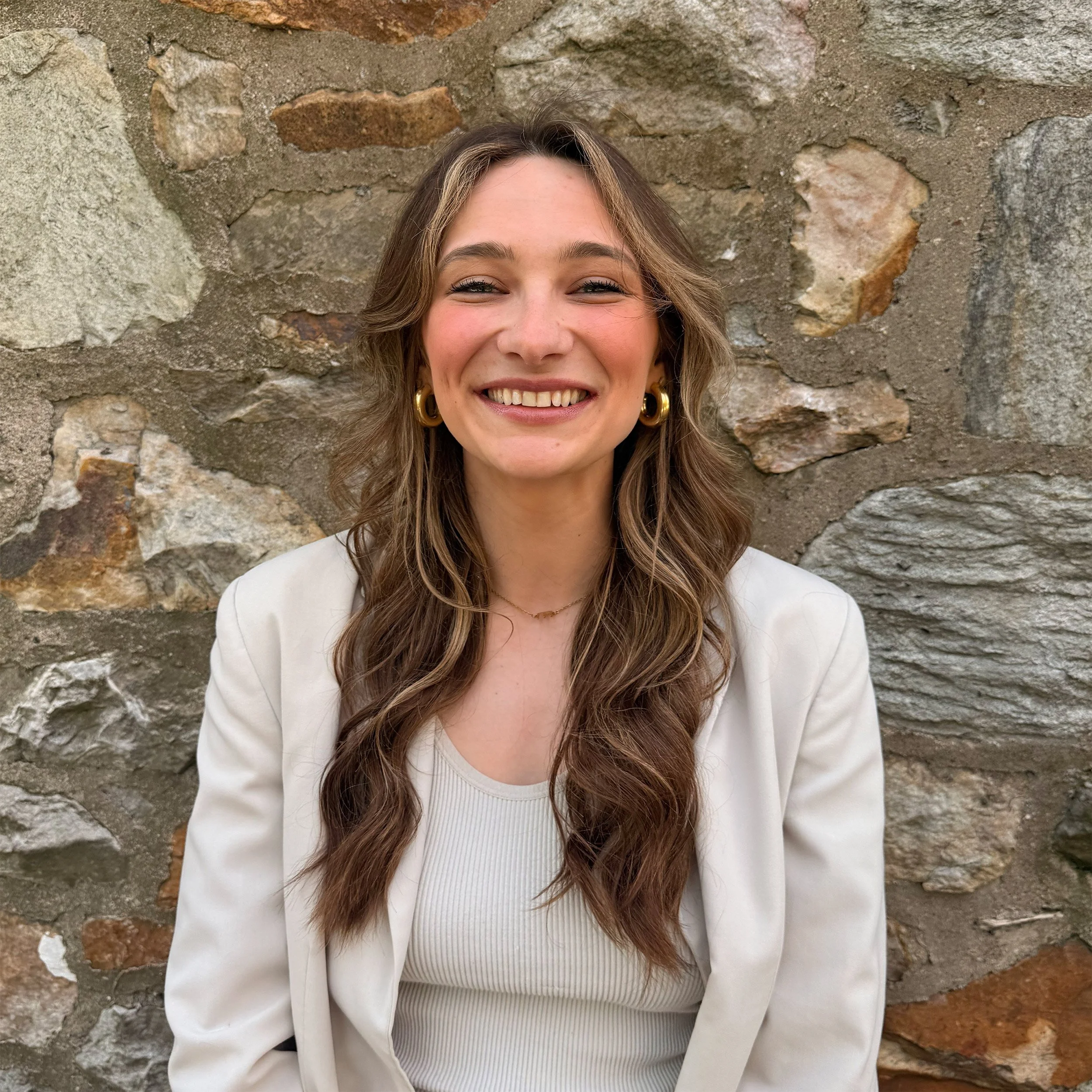 Smiling woman with long, wavy brown hair wearing gold hoop earrings, a white blazer, and a white ribbed top, standing in front of a rustic stone wall.
