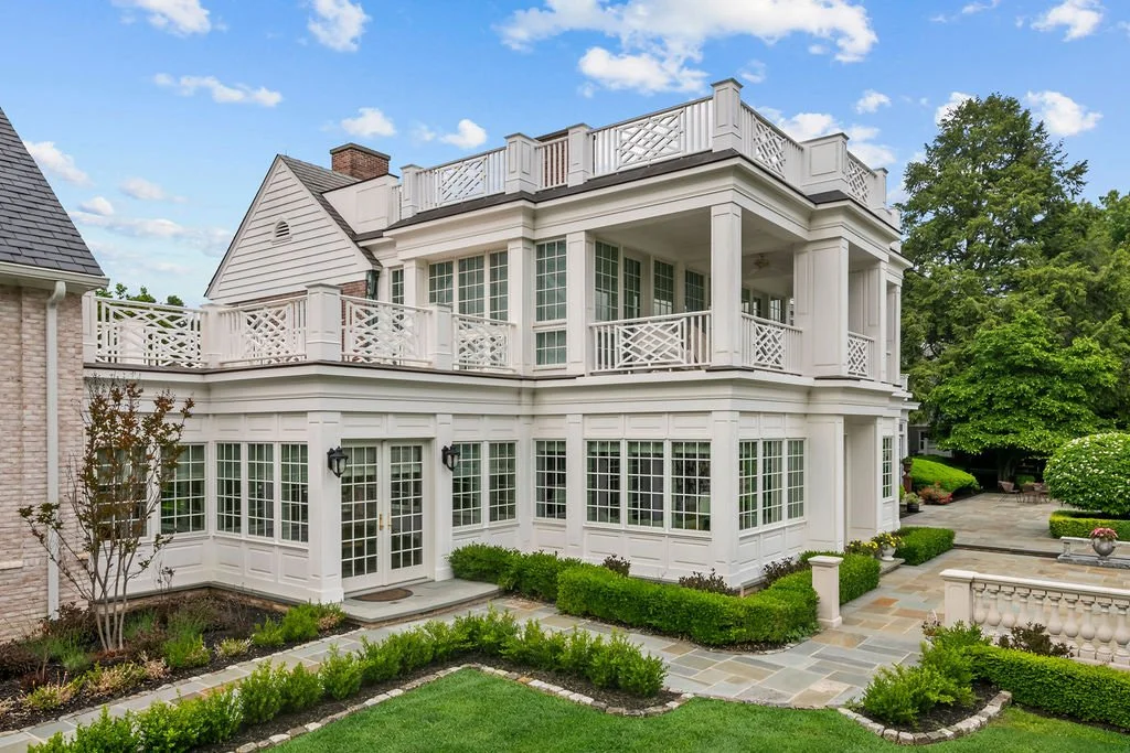 Large white multi-story house with wraparound balconies and multiple windows, surrounded by landscaped garden with bushes, trees, and paved walkways.