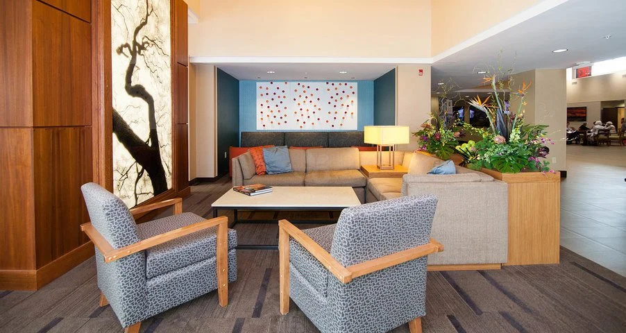Hotel lobby seating area with beige sectional sofa, two light blue patterned armchairs, a wooden coffee table with magazines, a lamp, and a large flower arrangement. Decor includes a artwork with dots and a tree mural.