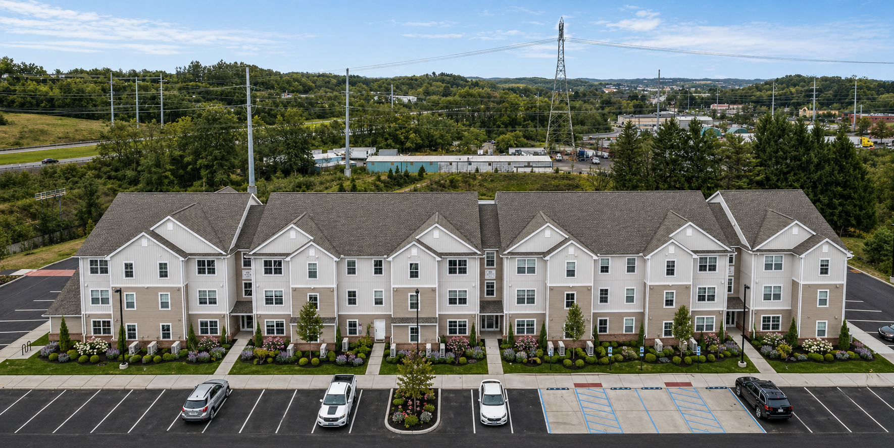 A row of modern apartment buildings with well-maintained landscaping, parking spaces, and cars parked in front. In the background, there are trees, power lines, and industrial buildings under a partly cloudy sky.