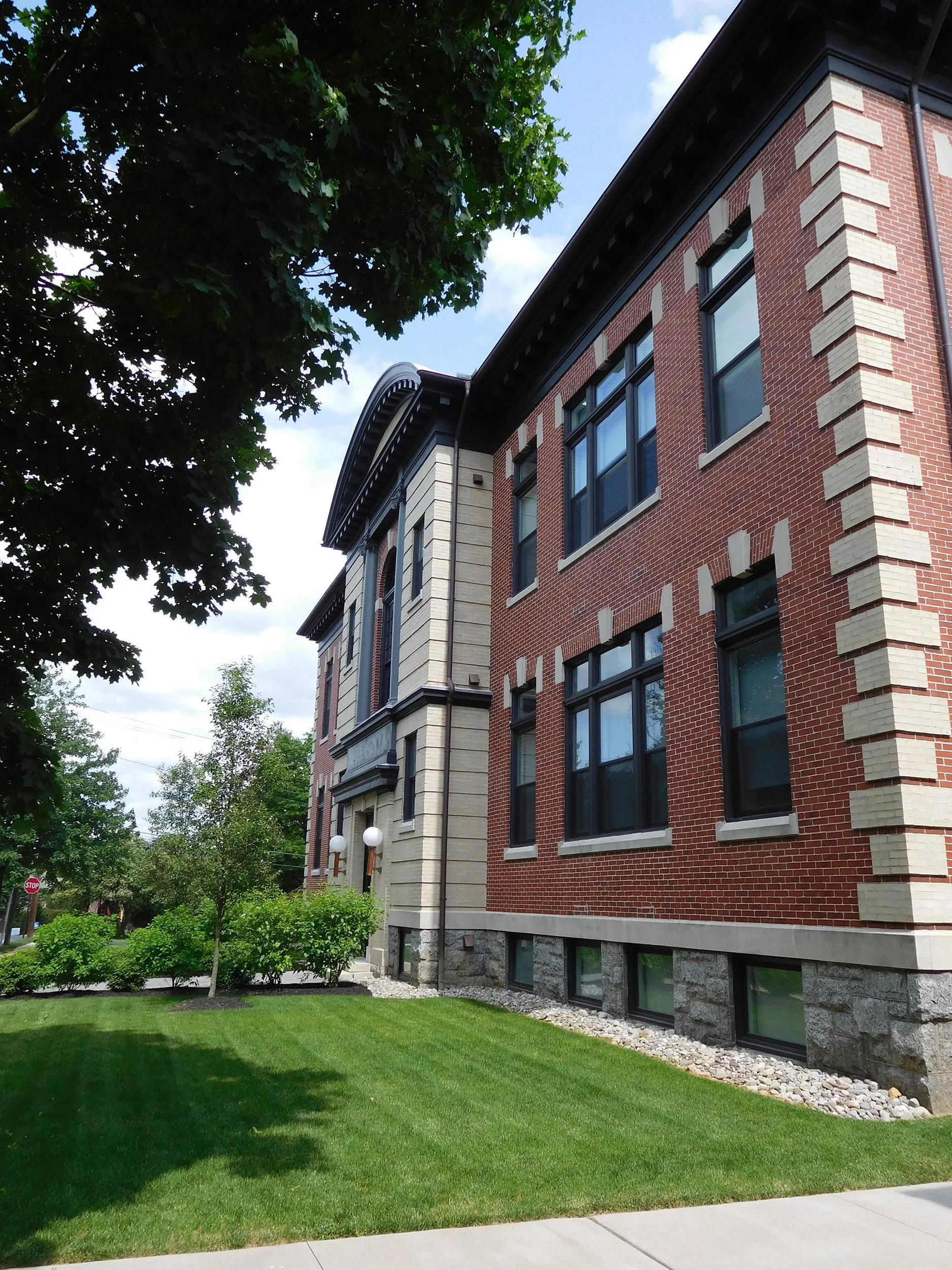 A multi-story brick apartment building with large windows and decorative stone accents. There is a well-maintained grassy lawn in front. A tree with green leaves partially frames the scene on the left, and the sky is partly cloudy.