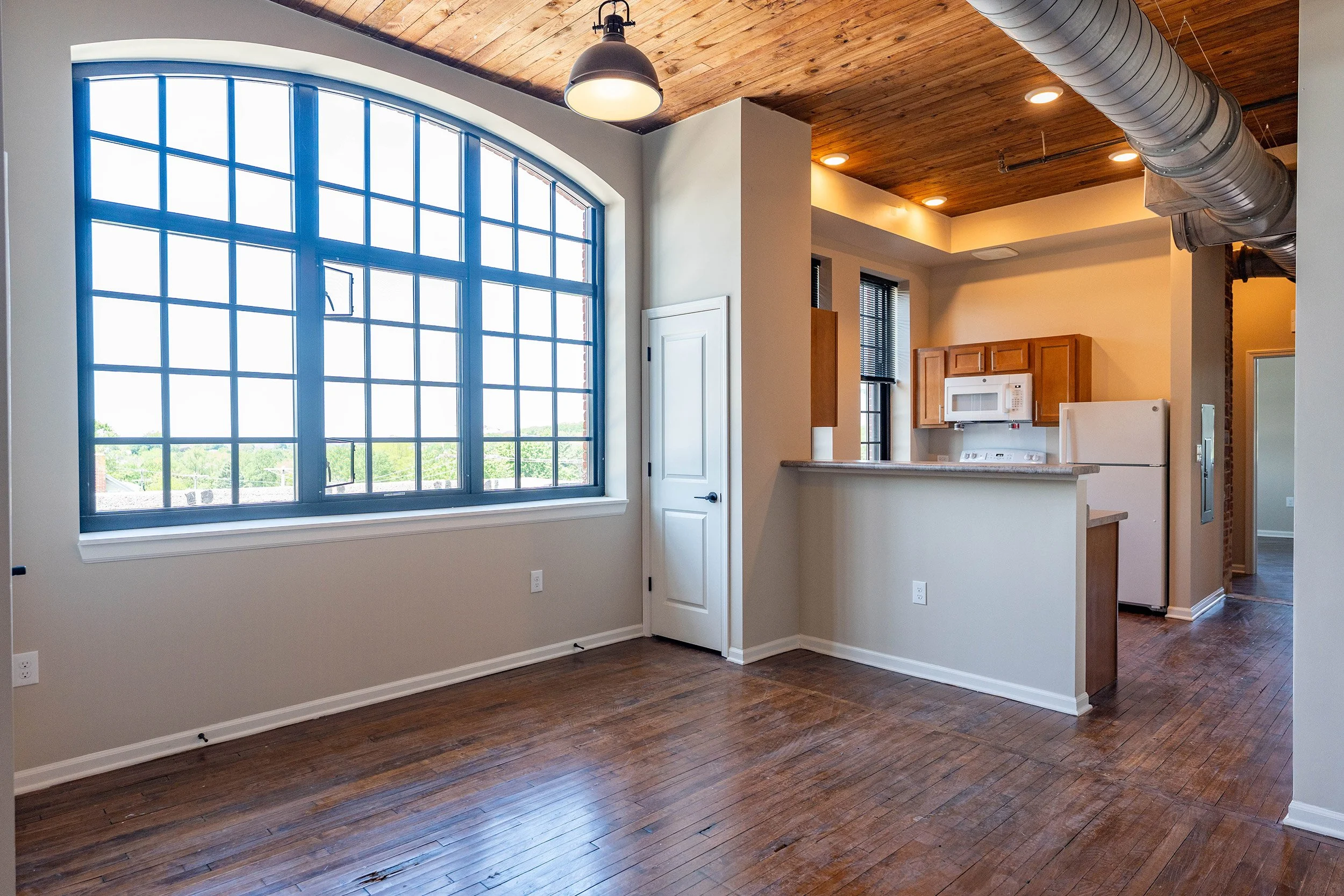 Empty apartment with a large arched window, hardwood floors, and a kitchen in the background featuring wooden cabinets, a white microwave, and a white refrigerator.