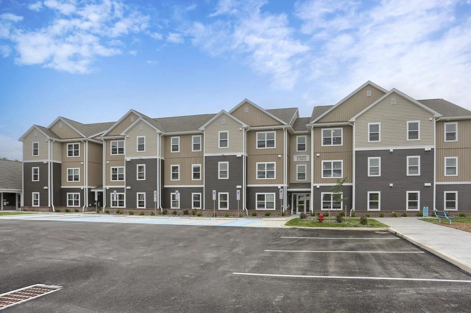 Exterior view of a multi-story apartment complex with beige and gray siding, multiple windows, a parking lot with marked spaces, a sidewalk, and a small landscaped area with shrubs and flowers, under a partly cloudy sky.