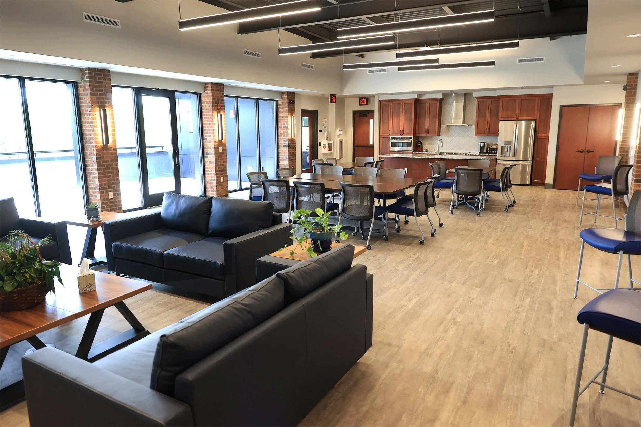 Modern lounge area with black leather sofas, wooden tables, and potted plants. In the background, there is a kitchen with wooden cabinets, stainless steel appliances, and a large dining table with chairs. Large windows with brick accents allow natura