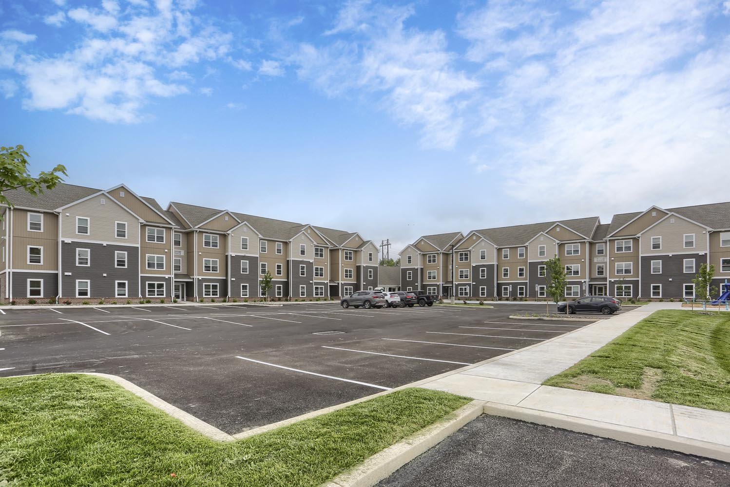 Multi-story apartment complex with a large parking lot in front, some cars parked, small trees lining the parking area, with a grassy area and sidewalk nearby, under a partly cloudy sky.