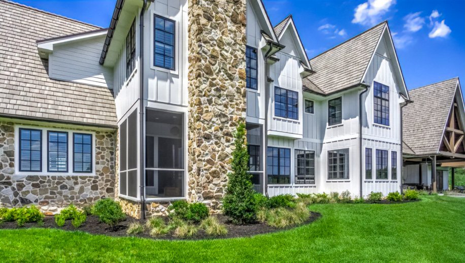 A modern house with stone and white siding exterior, multiple large windows with black frames, and a green lawn with landscaped bushes and small trees.