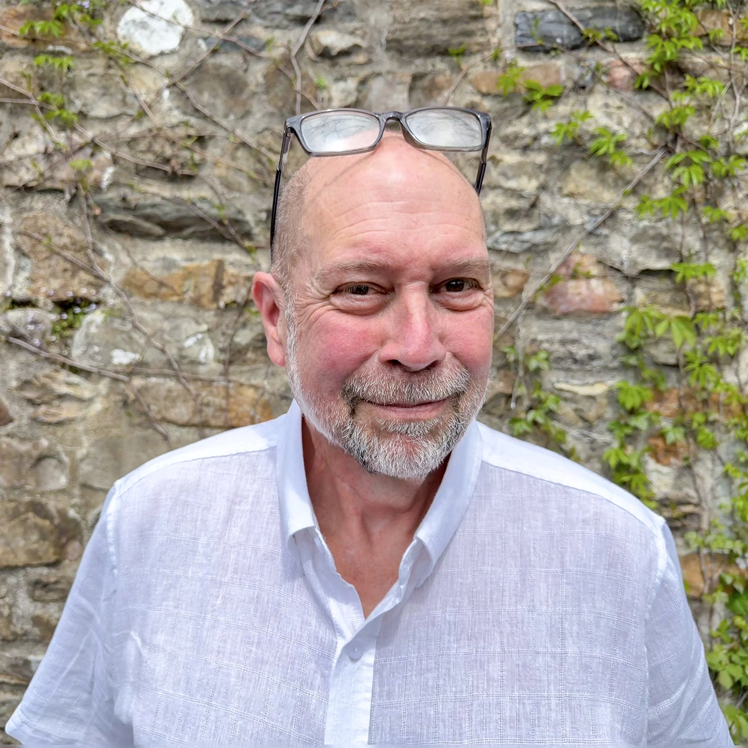 Close-up of a smiling man's face with a pair of eyeglasses resting on his bald head, standing in front of a stone wall with green vines.