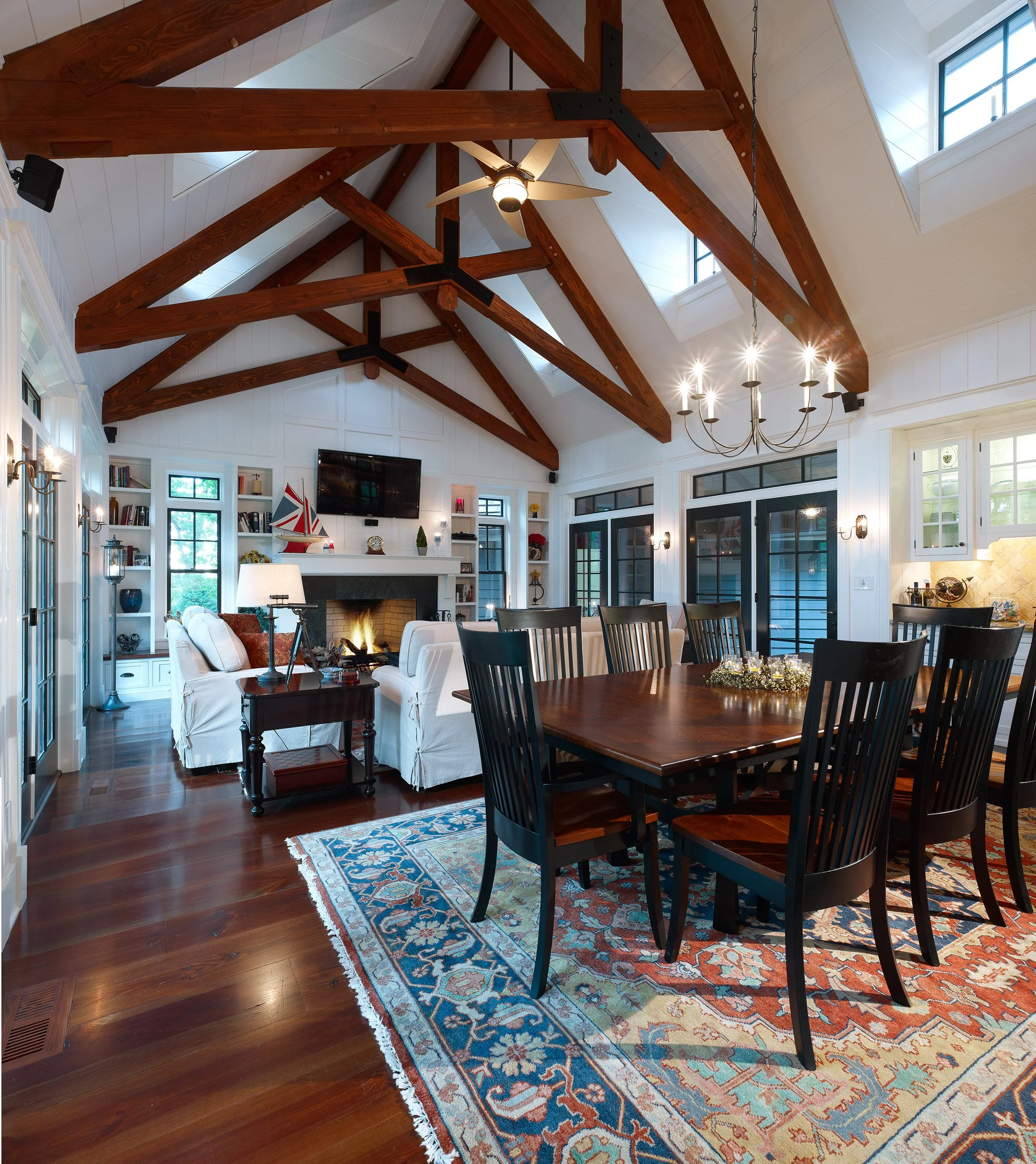 A spacious living and dining room with high vaulted ceiling, wooden beams, and windows in a modern farmhouse style. Features a fireplace, a mounted TV, and a dining table with black chairs. The room is decorated with a chandelier, bookshelves, and va