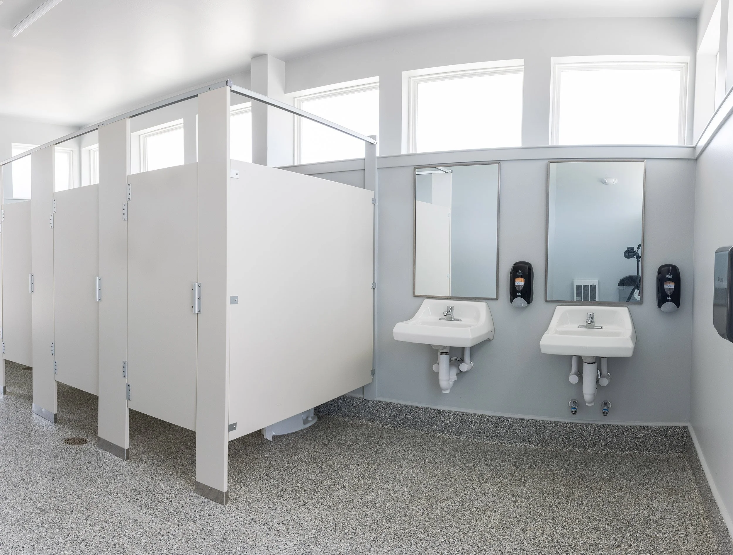Interior of an athletic complex restroom with two sinks, two mirrors, soap dispensers, hand dryers, and privacy stalls, with large windows letting in natural light.