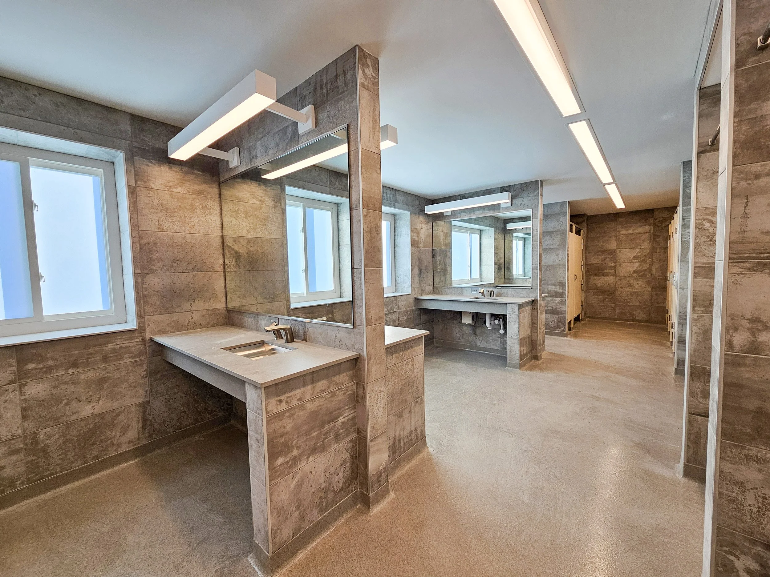 A modern dormitory bathroom with multiple sinks, large mirrors, beige tiled walls, and large windows.