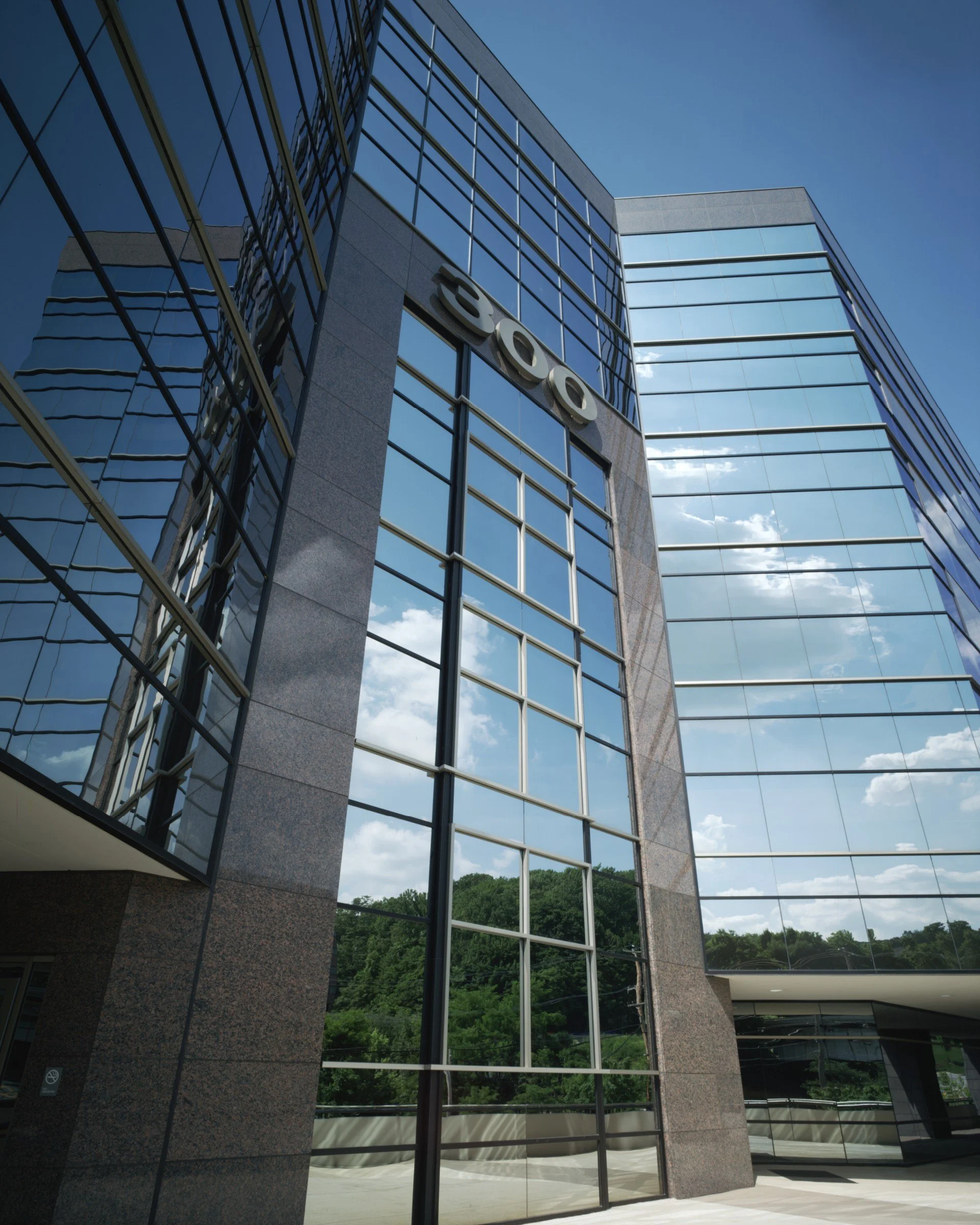 Modern office building with glass windows reflecting the blue sky and trees, with the number 300 displayed on the wall.