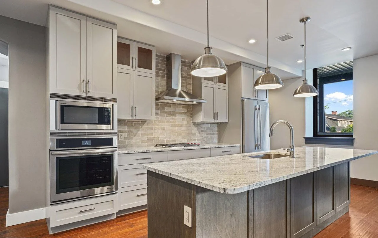 Modern kitchen with white cabinetry, stainless steel appliances, granite island, brick backsplash, pendant lighting, and hardwood floors.
