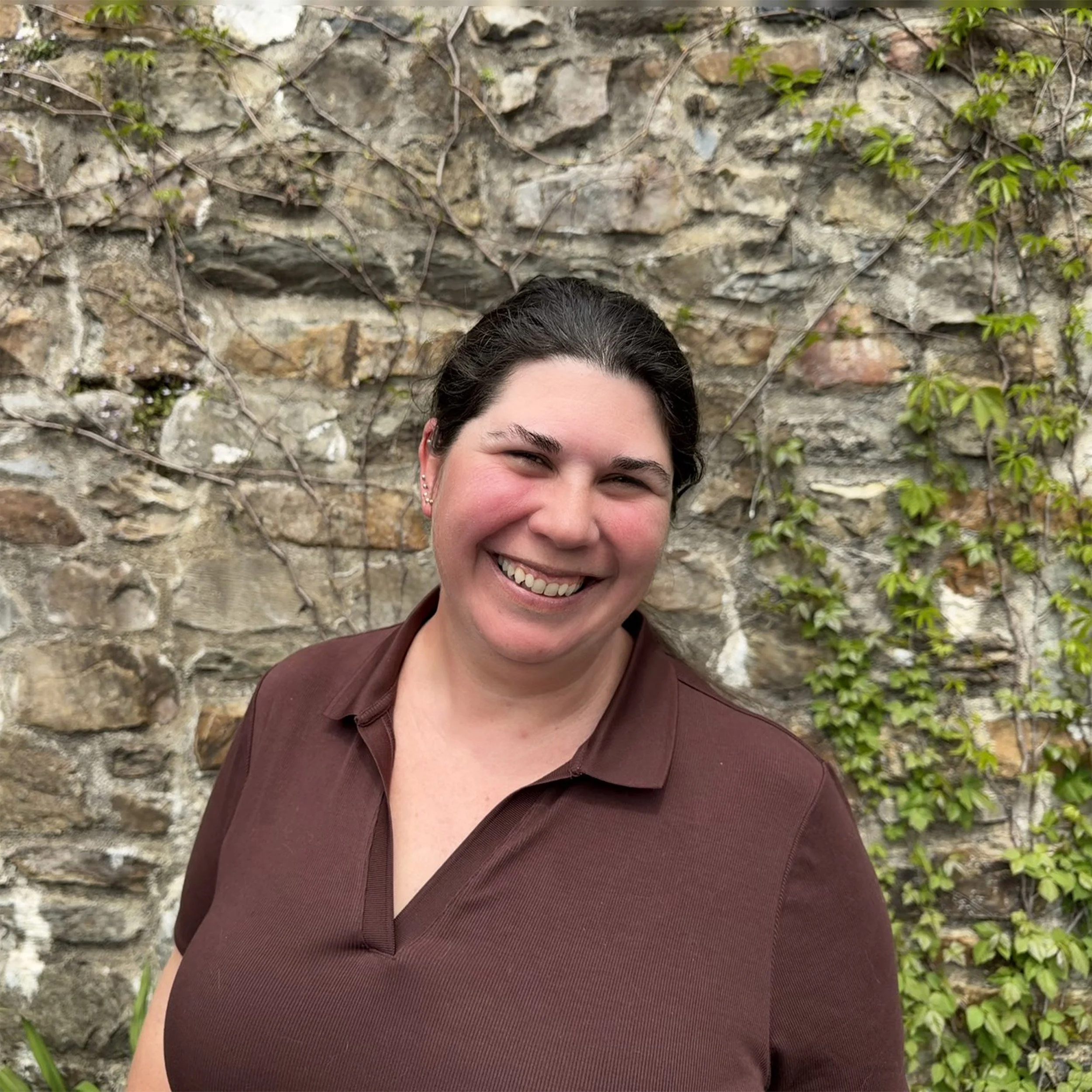 A smiling woman with dark hair and earrings standing in front of a stone wall with vines and green leaves.