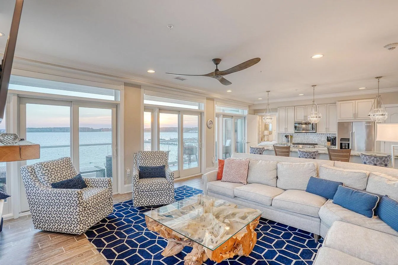 Living room with large windows overlooking a water view, featuring a beige sectional sofa with blue and pink pillows, two patterned armchairs, a glass and wood coffee table, and a blue geometric rug.