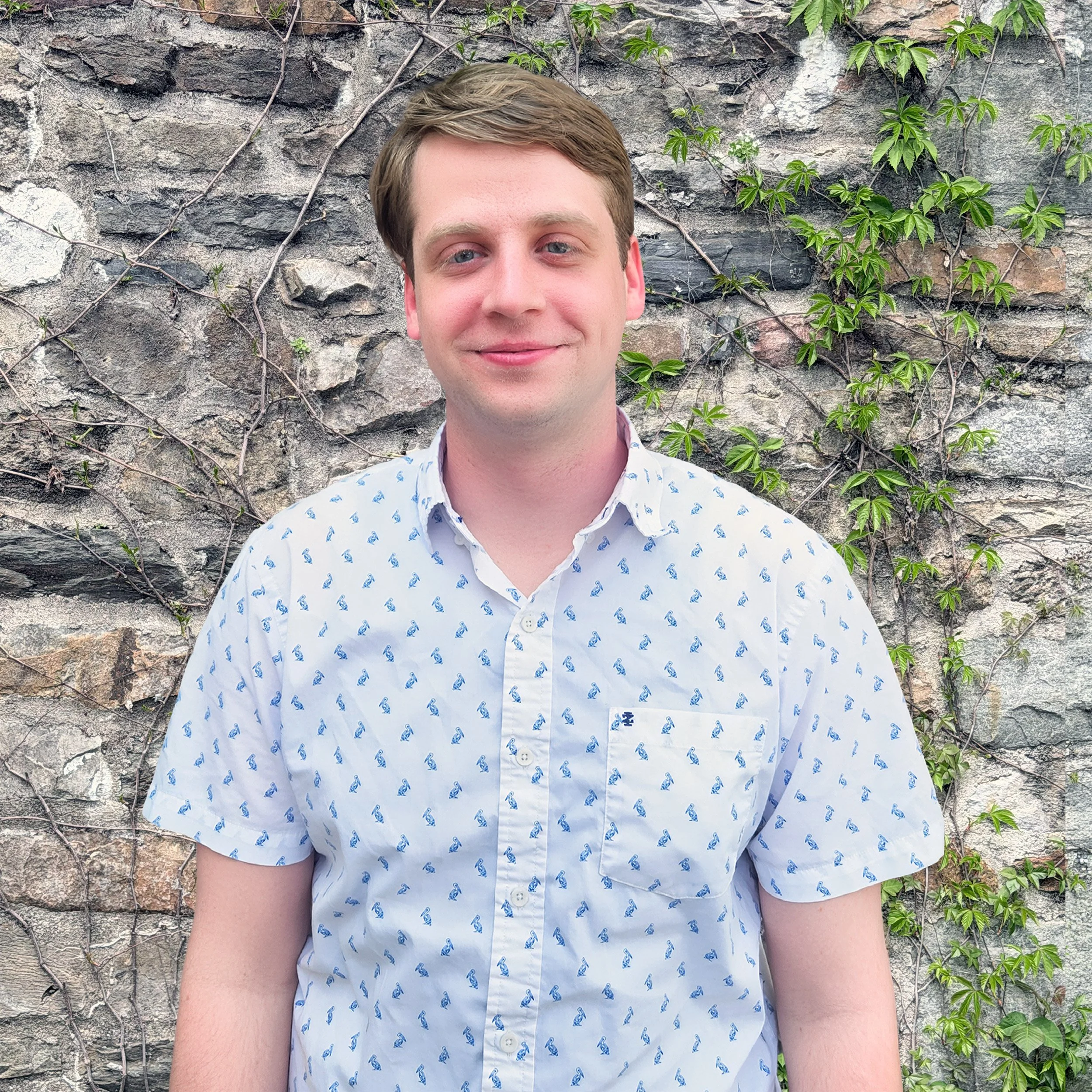 A young man with light brown hair and fair skin, wearing a light-colored shirt with a small pattern, is smiling slightly and standing in front of a stone wall covered with green vines.