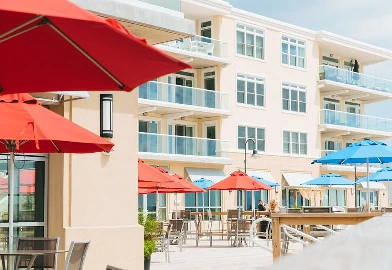 Outdoor patio area with red and blue umbrellas, tables, and chairs in front of a white multi-story building with balconies under a partly cloudy sky.