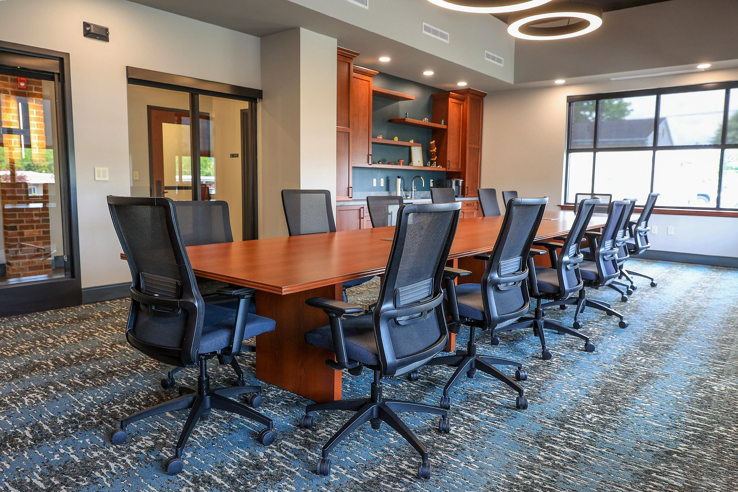 A conference room with a long wooden table and nine black office chairs on wheels. Large windows on one side, a kitchenette with wooden cabinets and a sink on the other, and a modern circular ceiling light.