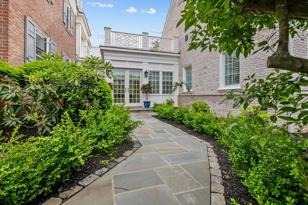 Stone pathway leading to the back of a house with white exterior and multiple windows, surrounded by lush green bushes and plants, with a tree on the right side and potted flowers near the entrance.
